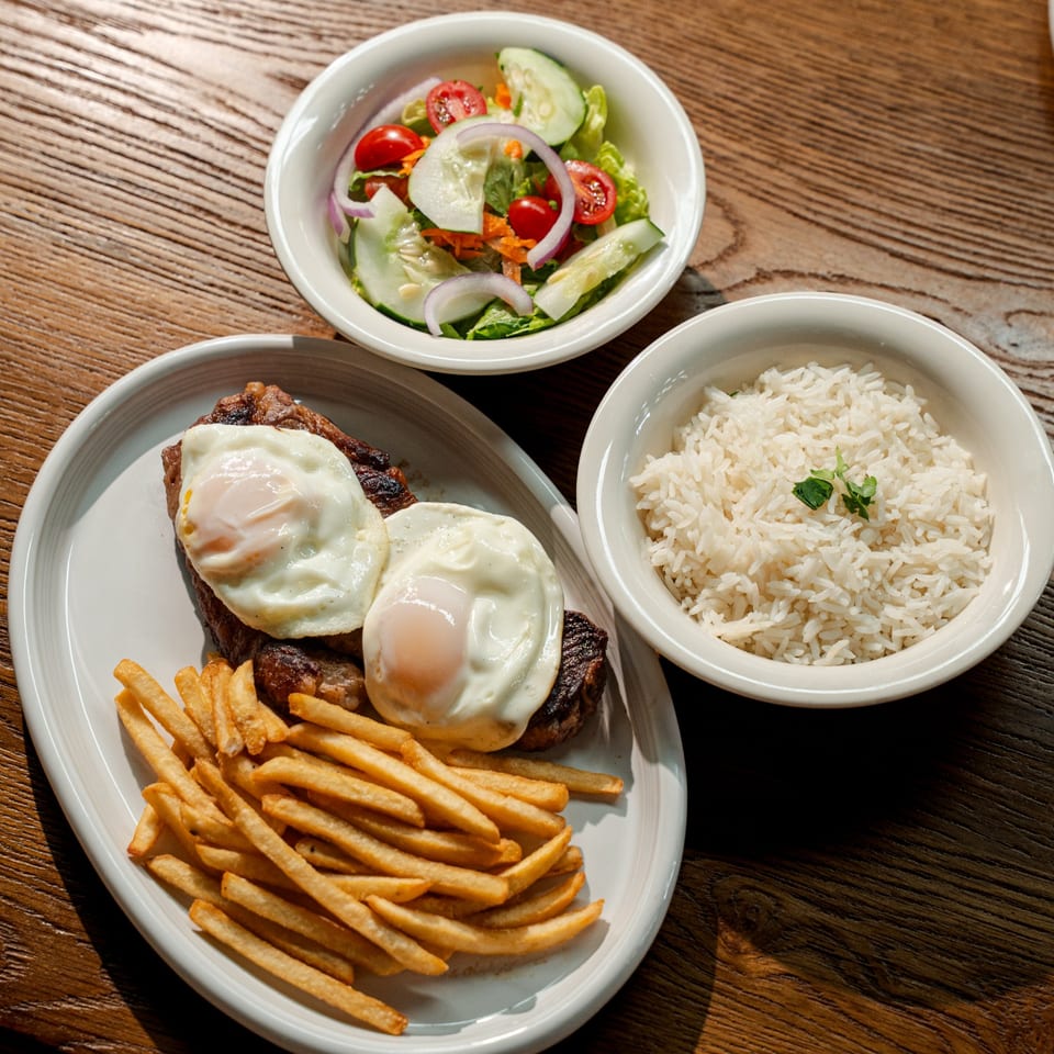 Grilled steak topped with two fried eggs, served with French fries, white rice, and a fresh cucumber-tomato salad.