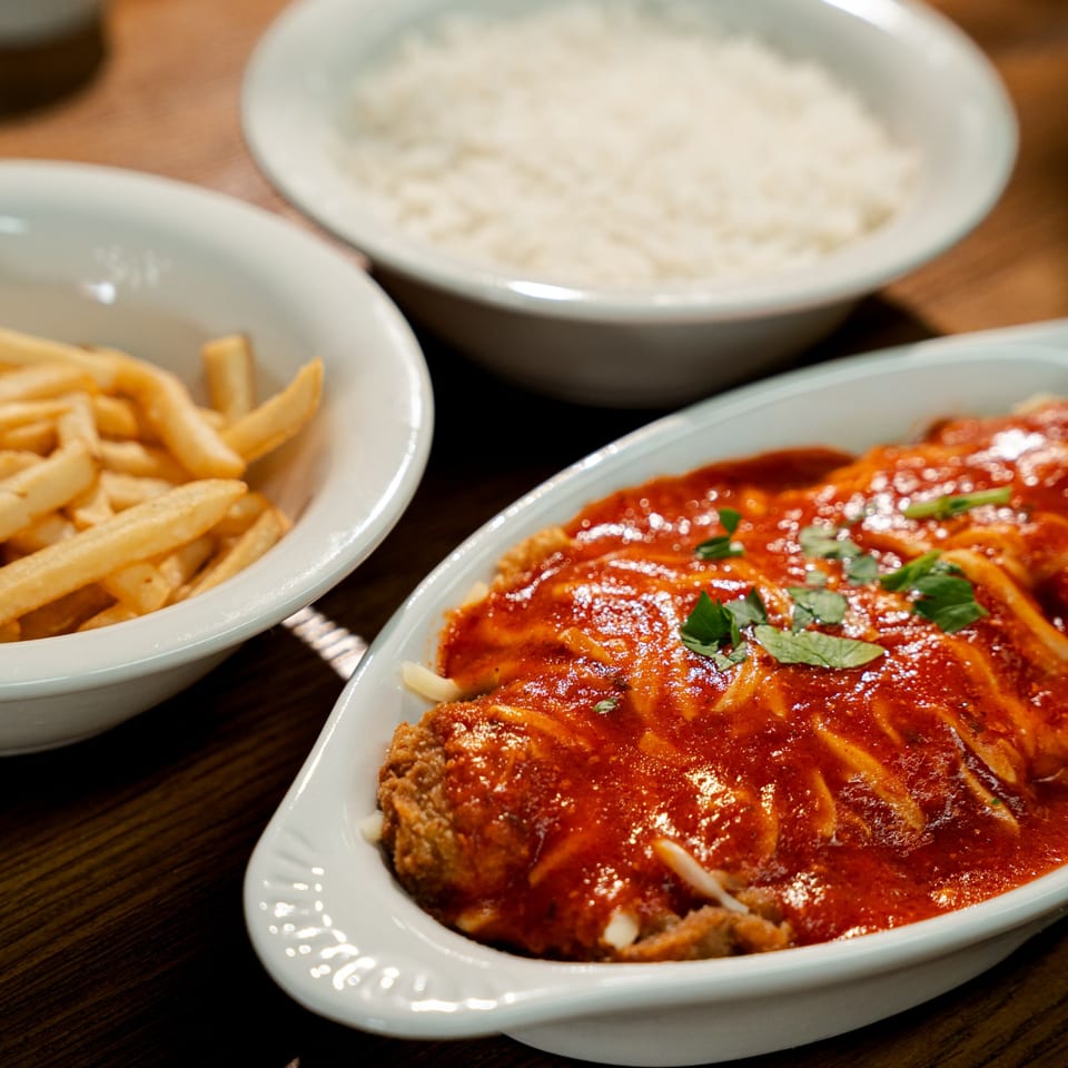 Breaded chicken covered in tomato sauce and herbs, served with fries and rice on a wooden table.