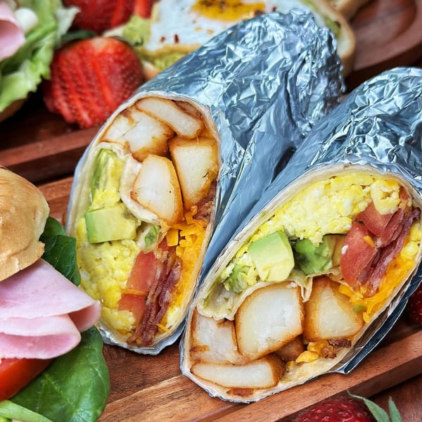 Two breakfast burritos, one cut open, revealing eggs, avocado, bacon, cheese, and potatoes.