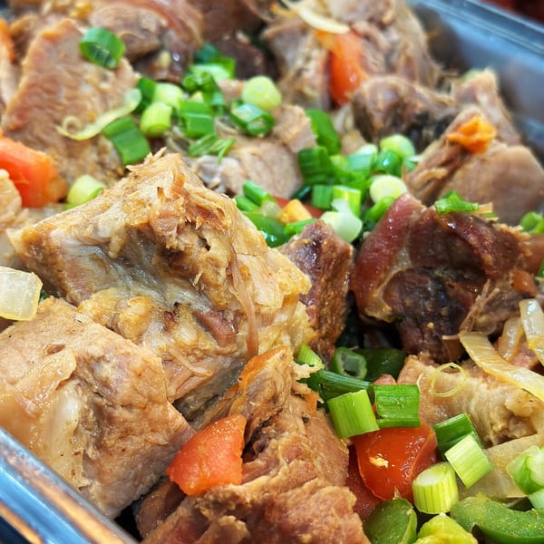 Close-up of Brazilian pork stew with green onions, tomatoes, and onions in a metal serving dish.