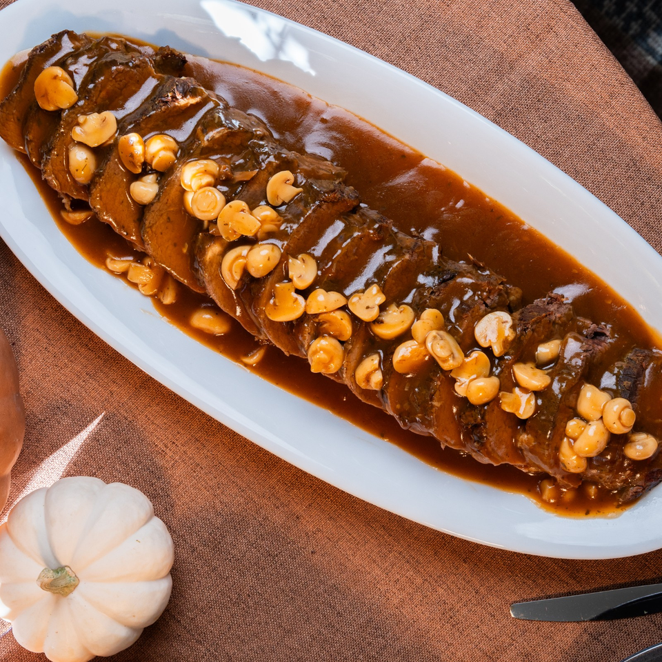 Sliced braised meat covered in glossy brown sauce and mushrooms on a white platter, with a decorative white pumpkin nearby.