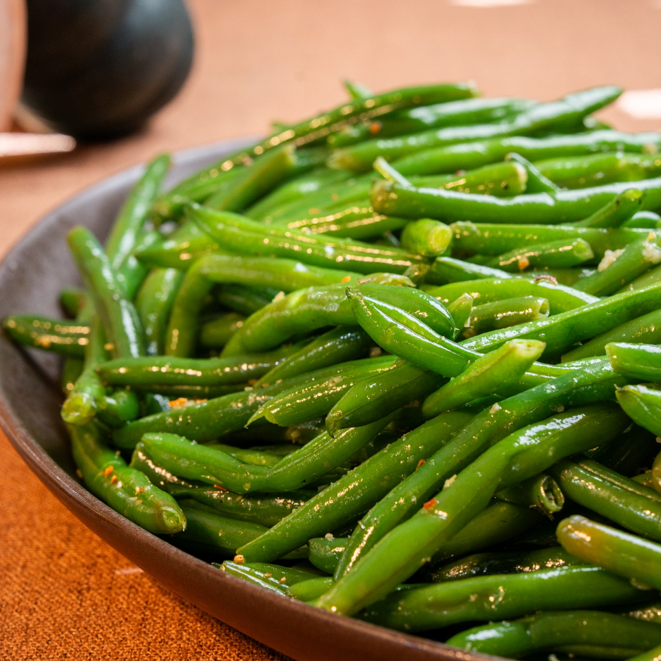 Glazed green beans seasoned with herbs and spices fill a dark serving bowl on a restaurant table.
